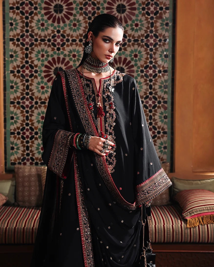 A woman models a traditional black embroidered kurta set with a red and gold border, styled with matching jewelry and a patterned tapestry in the background.