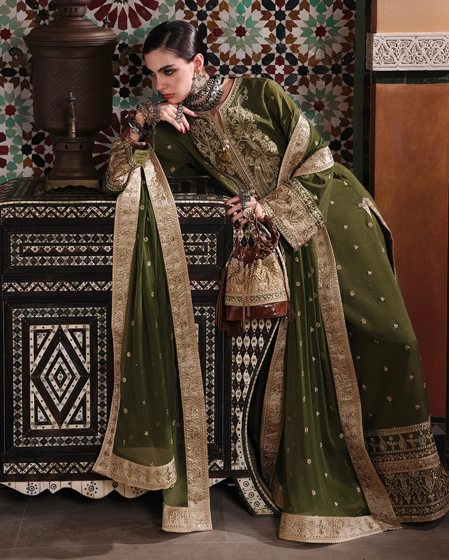 A woman models an olive green traditional South Asian outfit with intricate gold embroidery and a matching dupatta, posed in front of a patterned tile wall and a carved wooden cabinet.