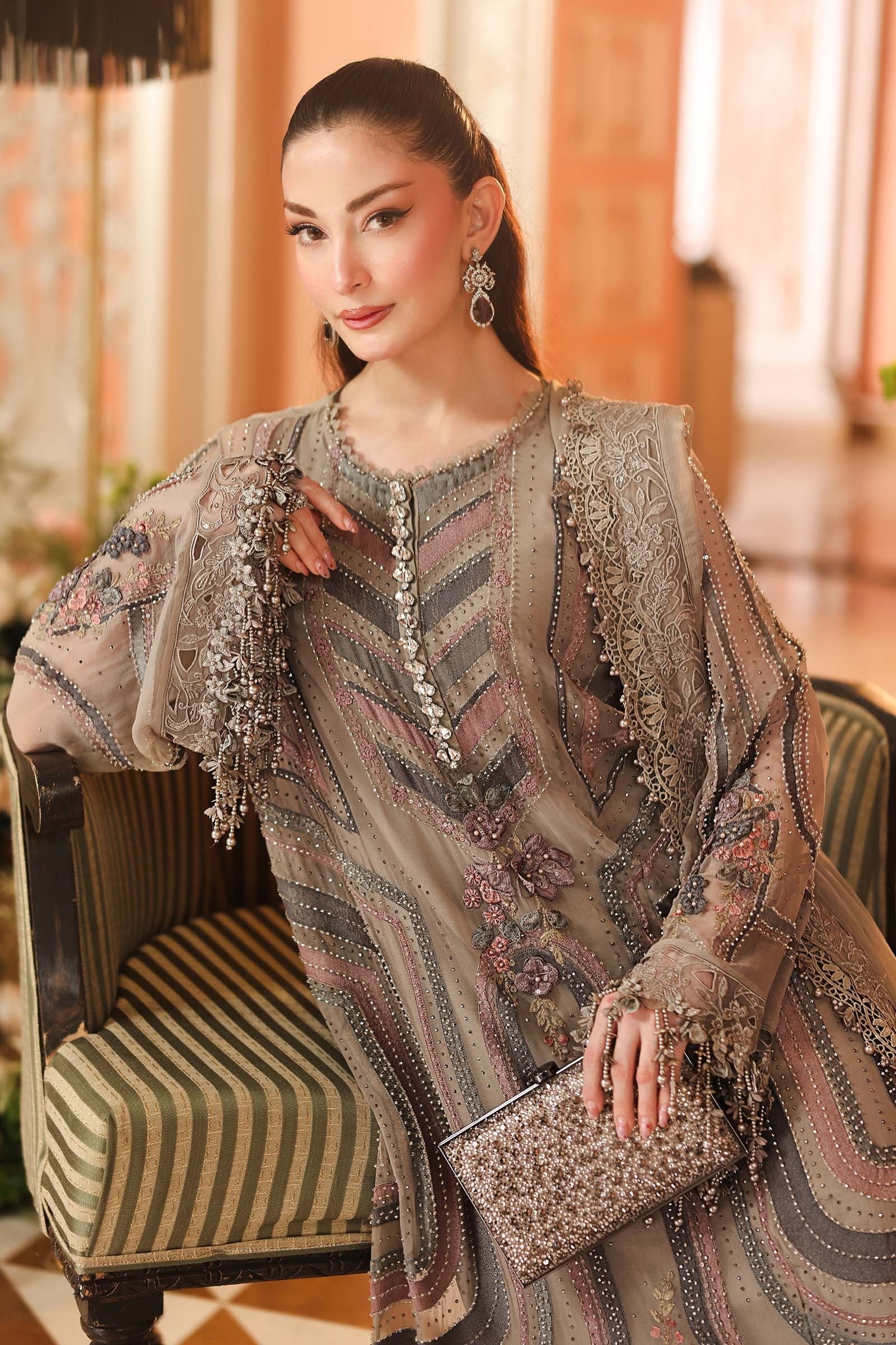 A woman models a traditional embroidered outfit in a warm indoor setting. The outfit is a sheer taupe-beige tunic with intricate floral embroidery, geometric patterns, and silver beadwork, paired with matching embellished sleeves and a matching clutch bag.