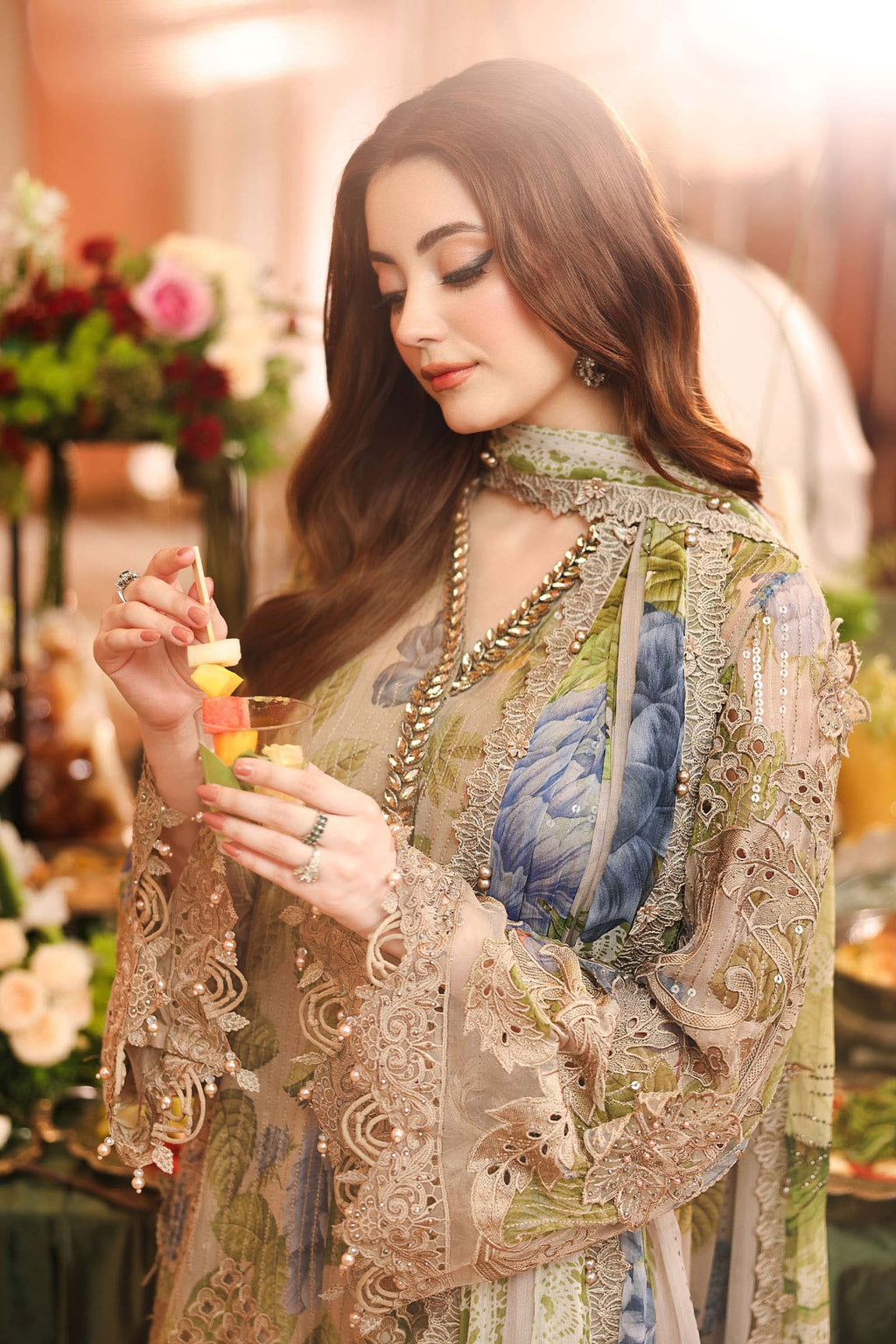 A woman in a detailed, ornate beige and blue printed traditional outfit with intricate lace embroidery, holding a small glass of water in a warm, decorated indoor setting.