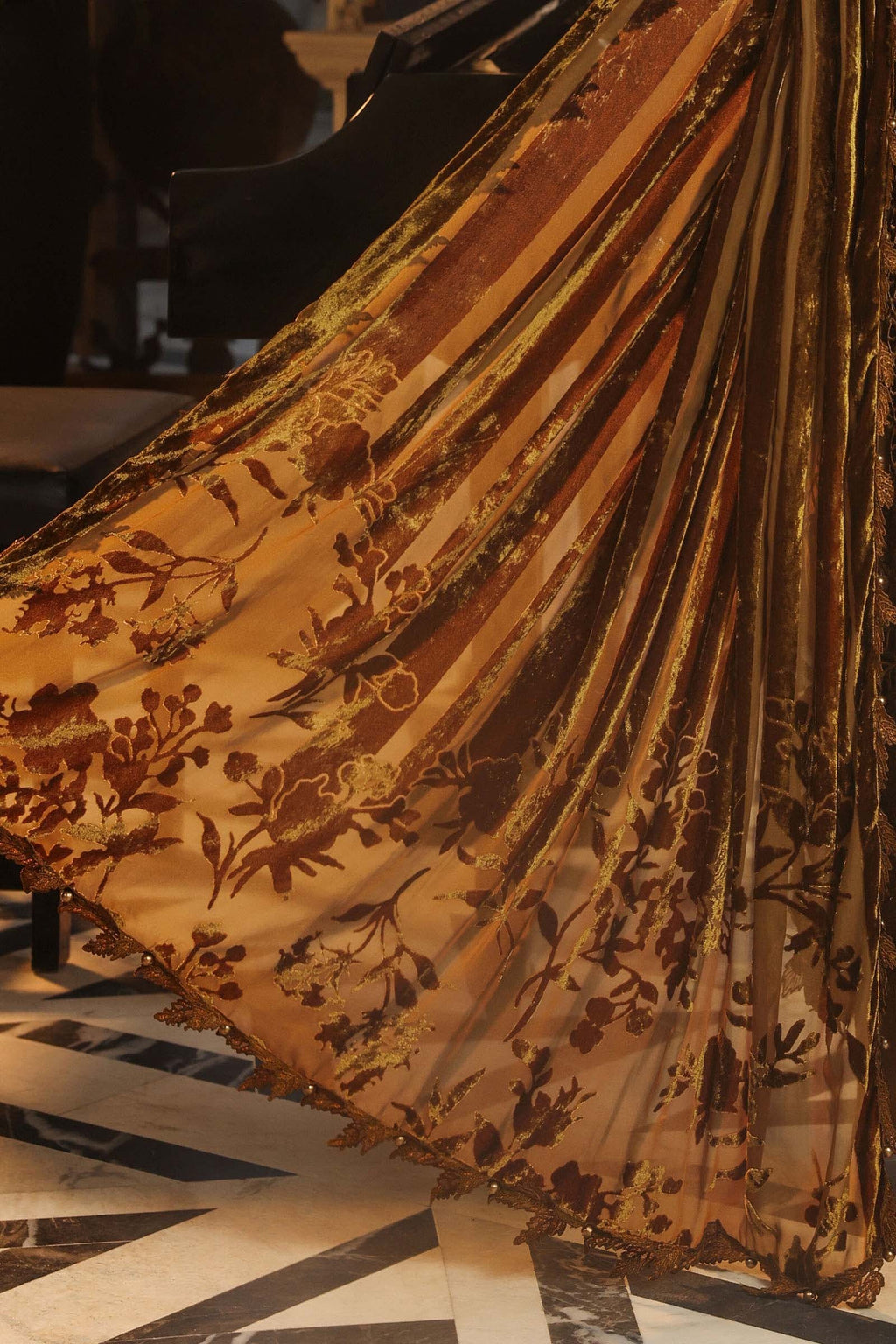 A close-up of a flowing sheer fabric with a warm tan and brown striped pattern and dark floral embroidery, draped dynamically over a piano in an indoor setting.
