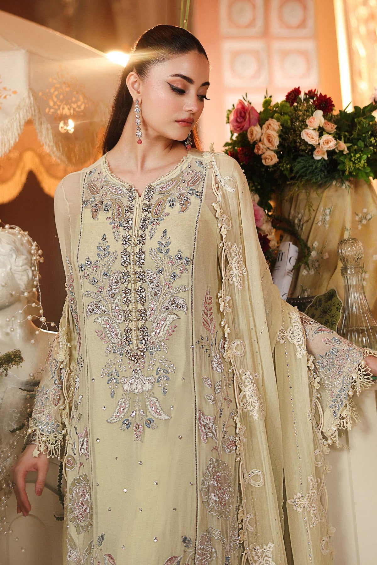A woman models a traditional South Asian salwar kameez set in a warm beige color with intricate silver, blue, and pink floral embroidery. The outfit includes a long kurta with a heavily embellished neckline and a matching sheer embroidered dupatta.