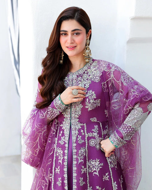A woman wearing a purple embroidered traditional outfit with sheer sleeves and silver floral detailing, photographed against a light architectural background.