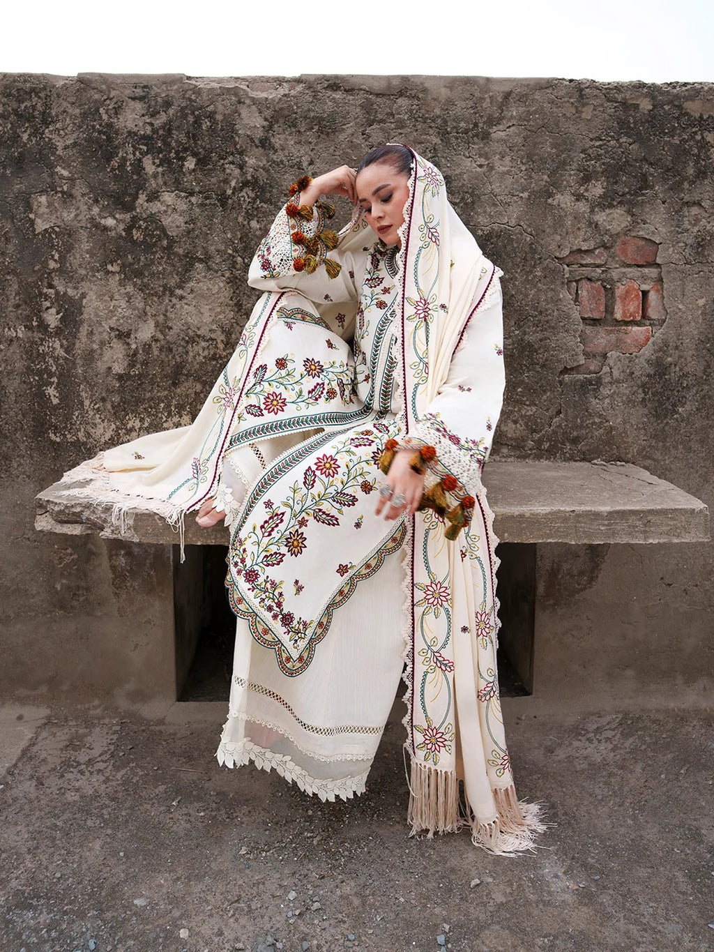 A woman sits on a rustic concrete bench against a weathered wall, wearing an ornate cream-colored embroidered robe and matching head covering with red, green, and blue floral patterns. She holds small dried floral clusters.