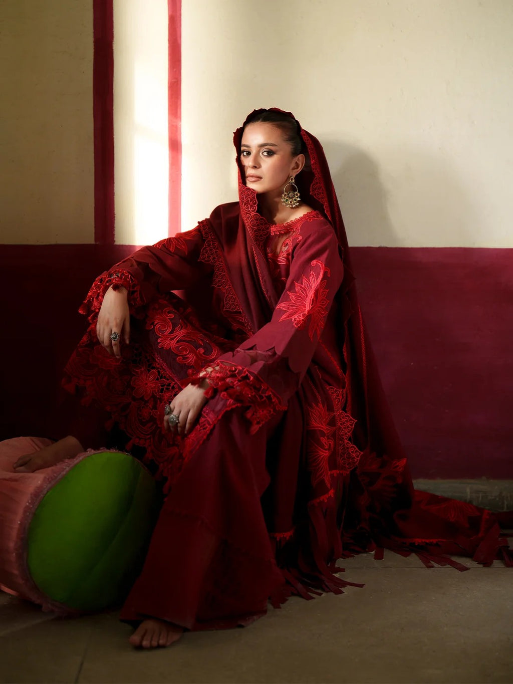 A woman seated indoors wearing an ornate deep red embroidered traditional outfit with a matching draped head covering. She is posed on a burgundy bench against a cream wall, with a green round cushion at her side.