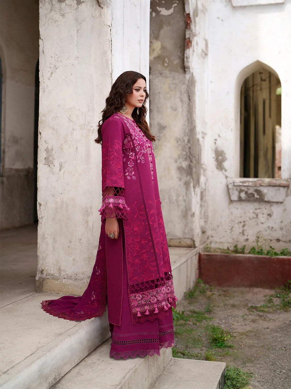 A woman wearing a long magenta-pink embroidered kurta and matching pants stands on stone steps in an outdoor courtyard with weathered white walls.