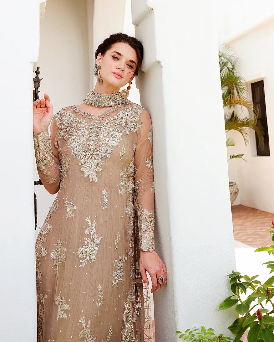 A woman models a beige embroidered traditional dress with sheer sleeves and intricate floral detailing, standing in a white architectural setting with plants.