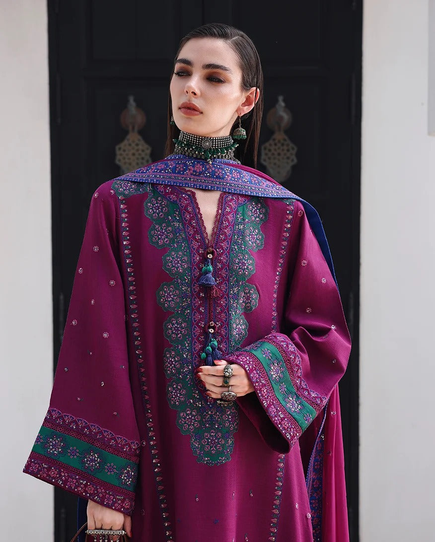 A woman models a traditional magenta-purple embroidered kaftan with green and blue patterns, a matching embroidered collar, and a dark blue shawl, set against a dark wooden door background.