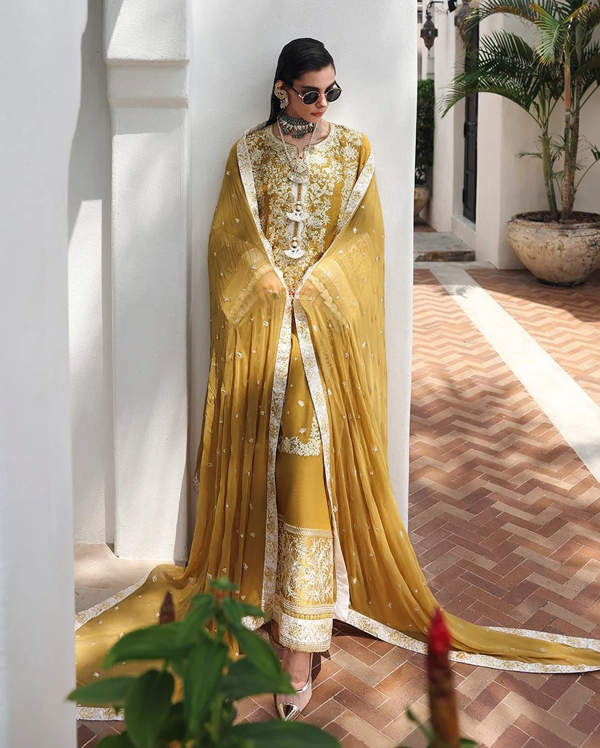 A woman models a traditional South Asian ensemble featuring a mustard yellow embroidered kurta and matching trousers, paired with a sheer matching dupatta draped over a white architectural column in an outdoor courtyard.