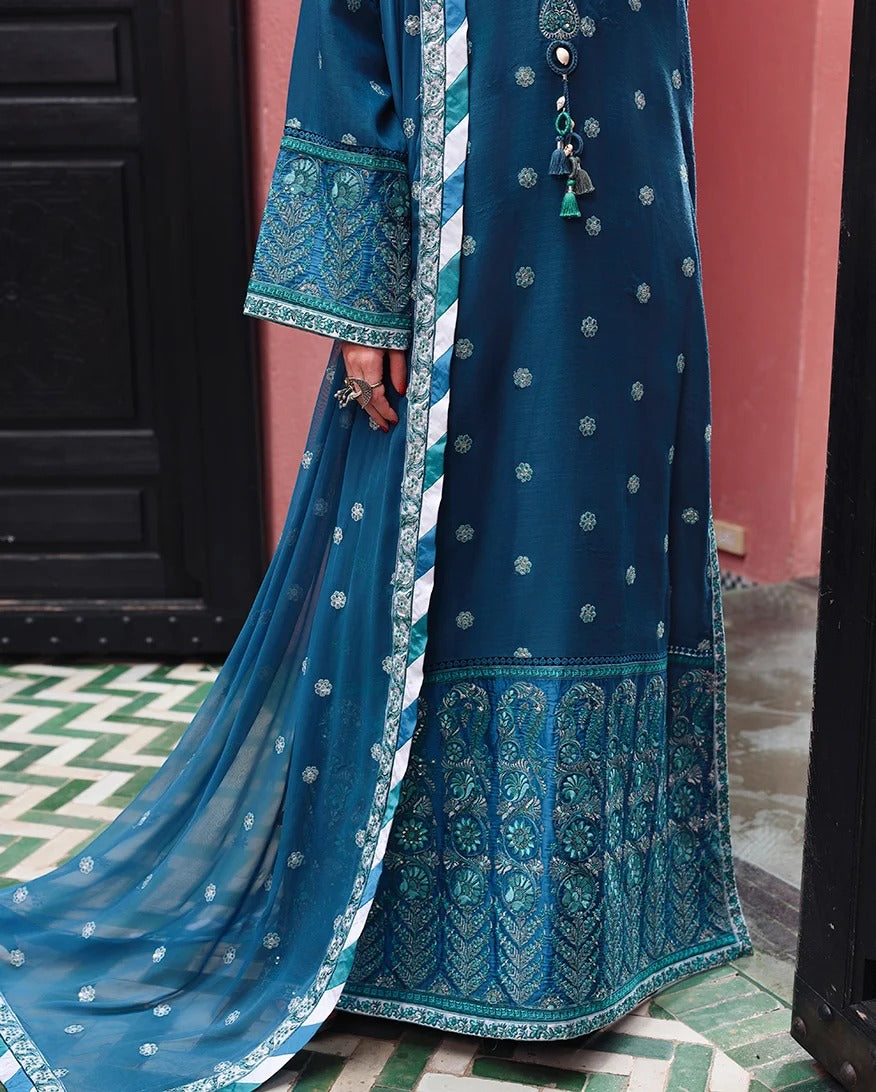 A close-up of a person wearing a traditional teal blue embroidered dress with a matching dupatta, featuring intricate silver and white floral patterns and a heavily decorated border, set against a pink wall and dark doorway.