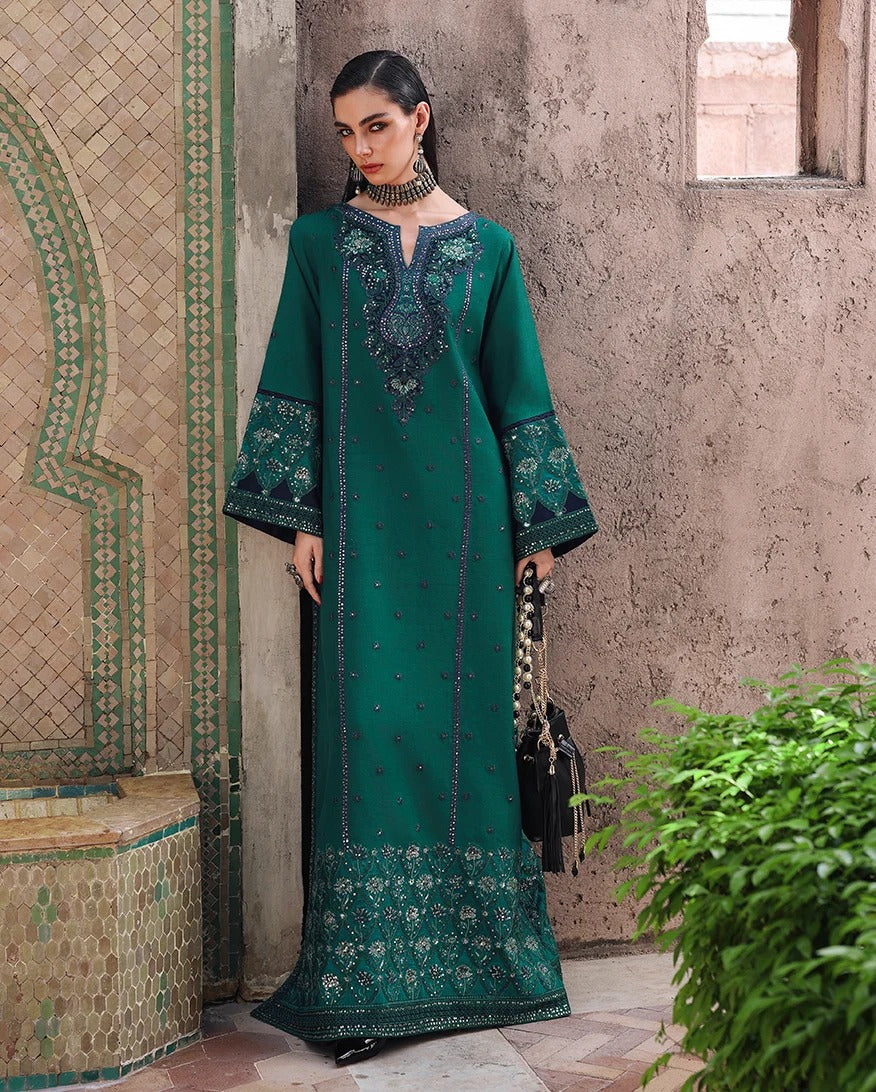 A woman models a long teal green kurta with intricate dark blue and gold embroidery, standing in a courtyard with a textured beige wall and tiled mosaic details.