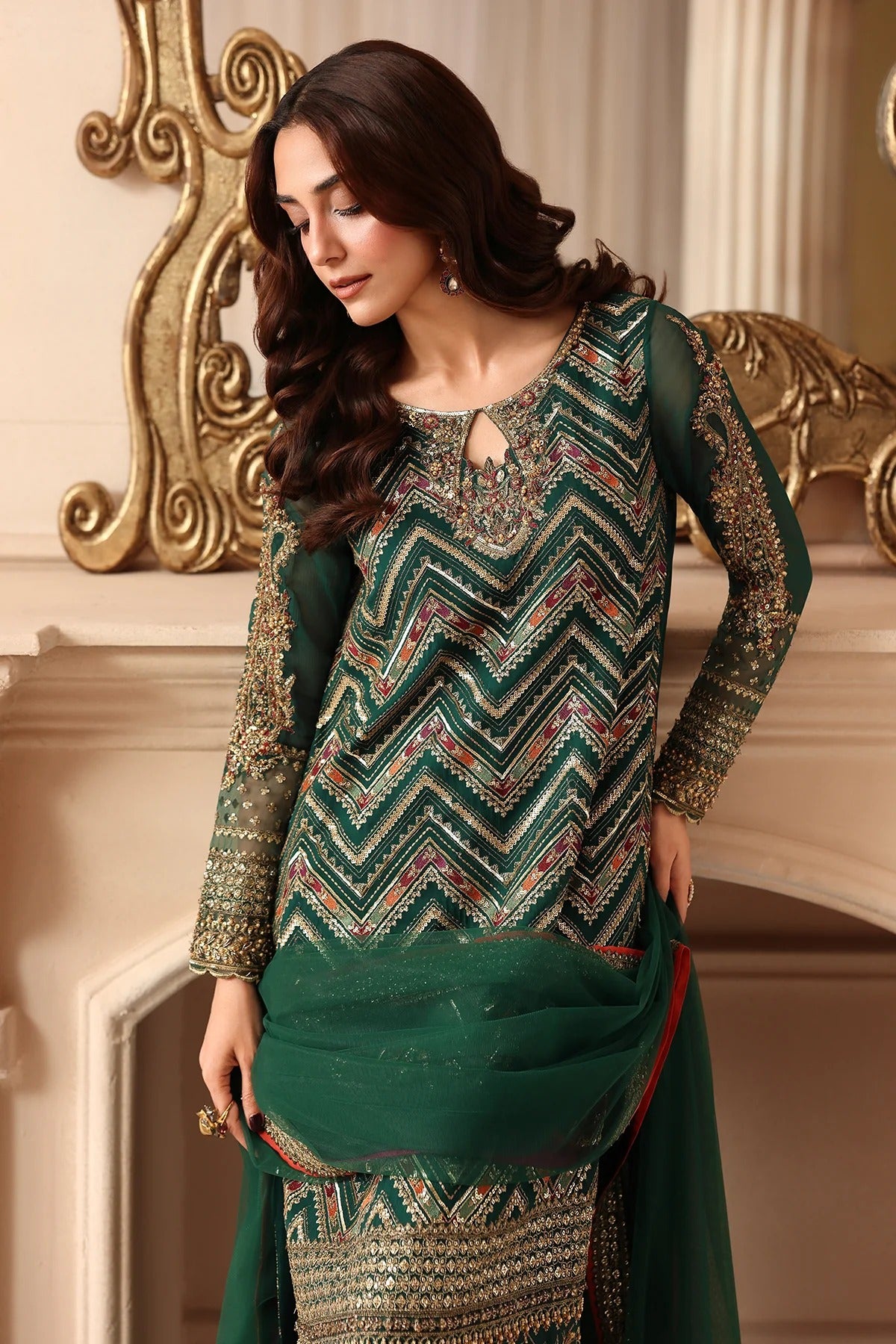 A woman models a dark green ethnic salwar kameez suit with intricate gold, silver, and red geometric embroidery, paired with a matching green dupatta, set against an ornate classical interior background.