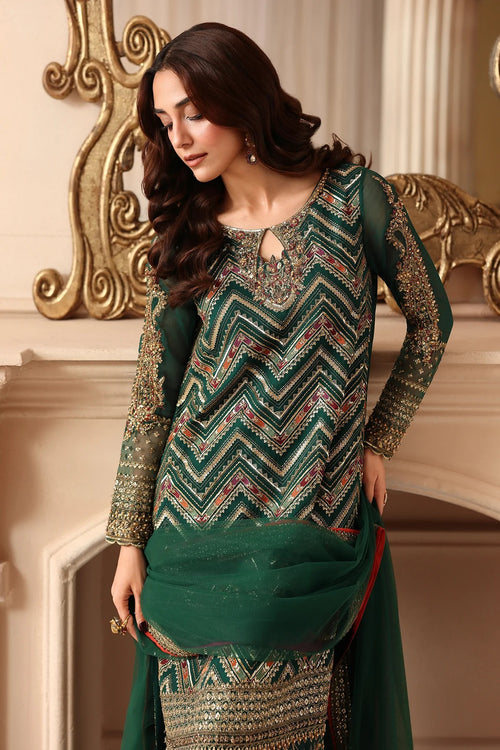A woman models a dark green ethnic salwar kameez suit with intricate gold, silver, and red geometric embroidery, paired with a matching green dupatta, set against an ornate classical interior background.