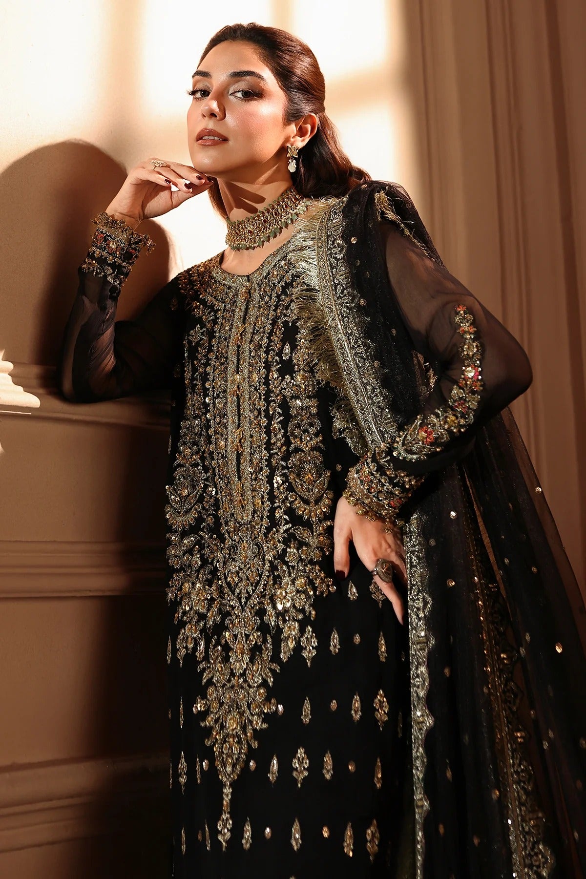 A woman models a black traditional embroidered outfit with ornate gold and silver embellishments, sheer sleeves, and a matching embroidered dupatta in a warm indoor setting.