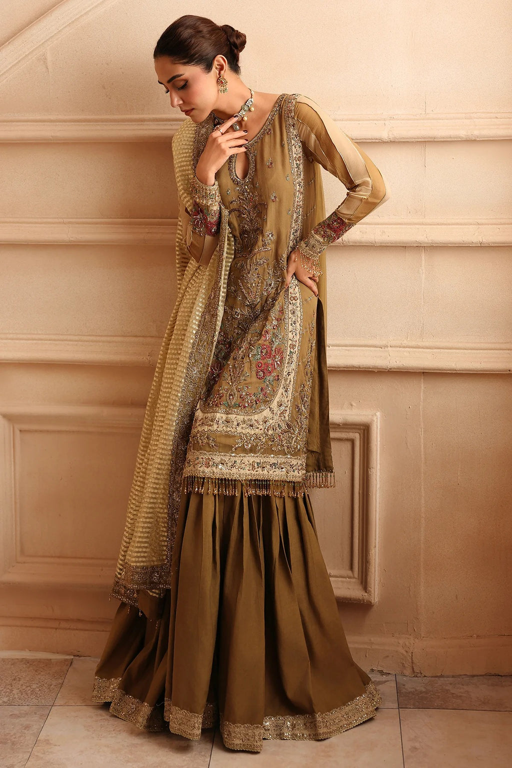 A woman models a traditional South Asian outfit consisting of a brown kurta with intricate gold embroidery, matching pleated skirt, and a sheer dupatta draped over her shoulder, set against a beige paneled wall.