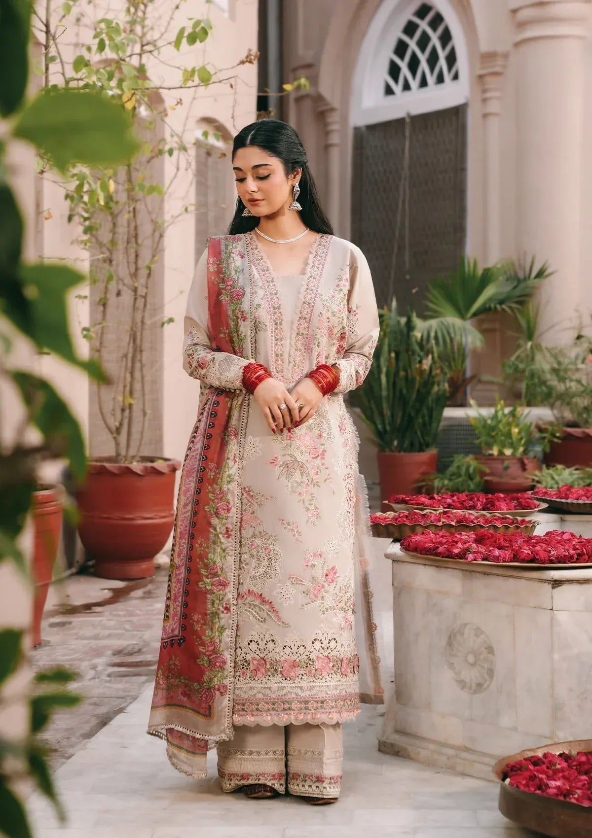 Woman in traditional outfit standing in a decorative outdoor setting with plants and flowers.