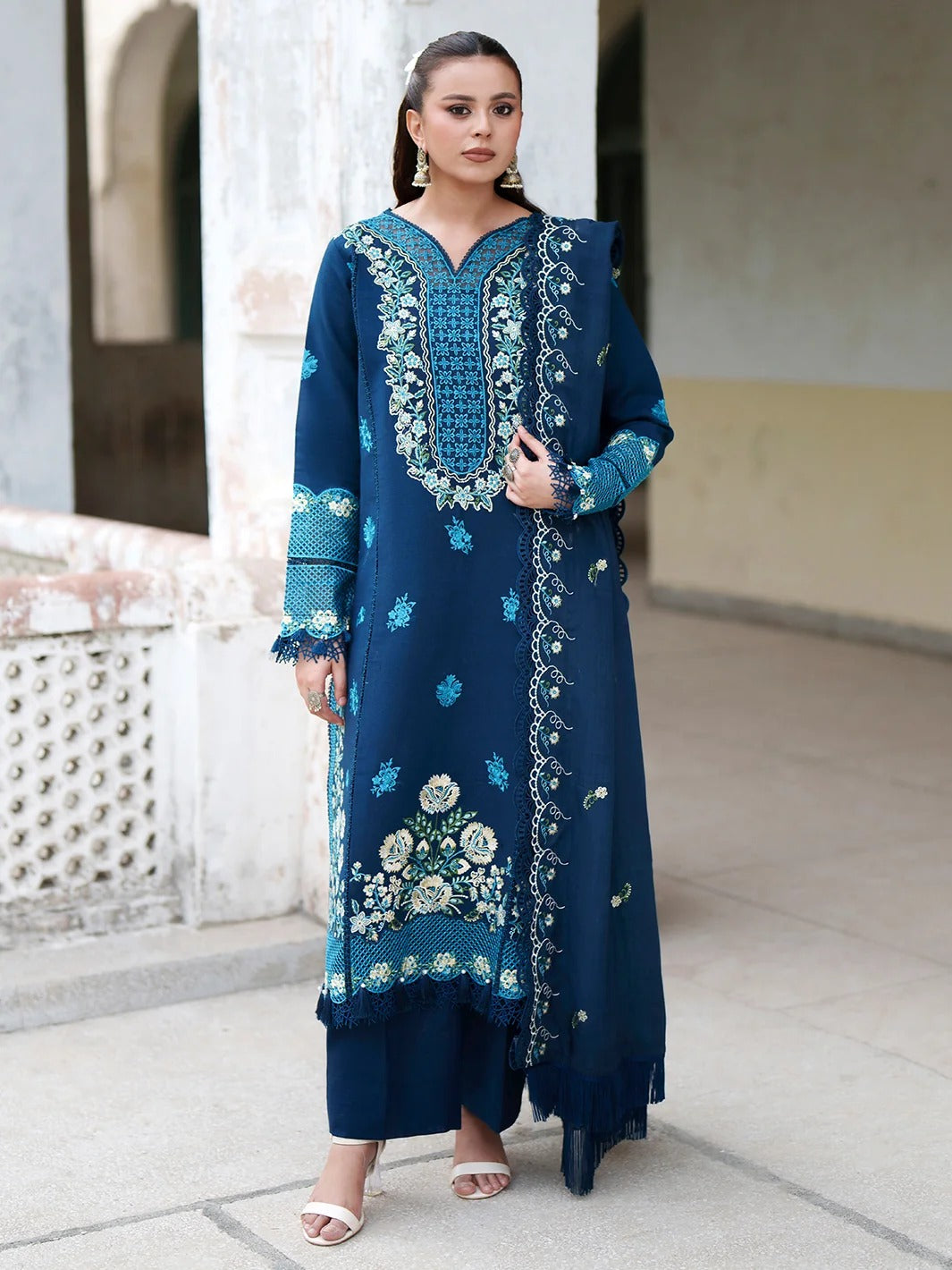 A woman modeling a dark teal blue traditional Indian salwar kameez suit with intricate light blue and white floral embroidery, standing in an outdoor architectural setting.