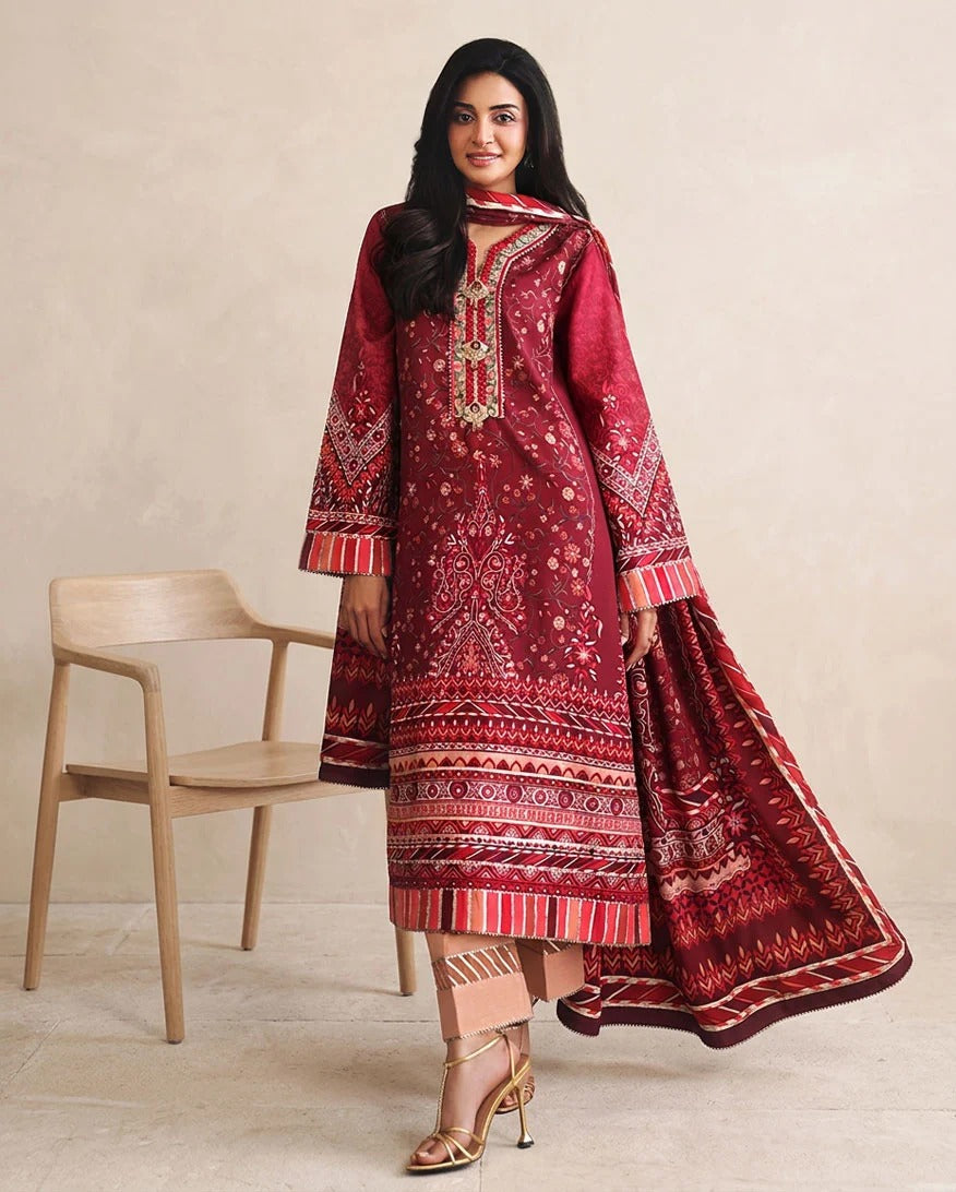 A woman models a traditional deep red three-piece salwar kameez suit with intricate gold and white embroidery, standing next to a light wood chair against a neutral beige background.