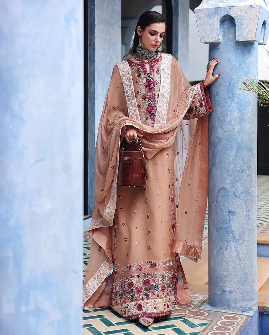 A woman models a traditional embroidered salwar kameez suit in a dusty peach-beige color, featuring intricate floral embroidery on the neckline, hem, and sleeves, paired with a matching dupatta and a brown leather handbag, set against a blue architectural background.