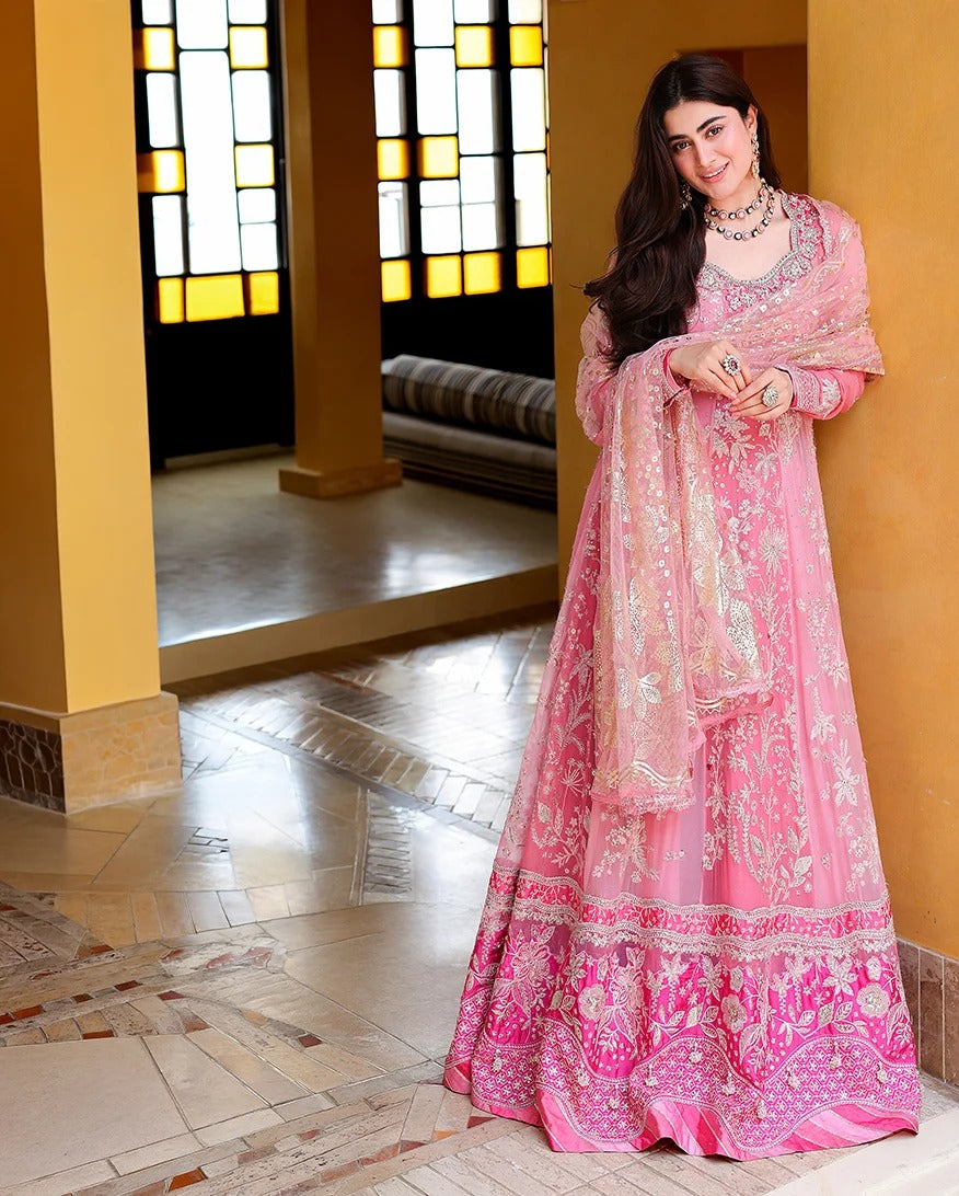 A woman models a pink embroidered traditional outfit with a matching dupatta in an indoor architectural setting with warm yellow walls and stained glass windows.