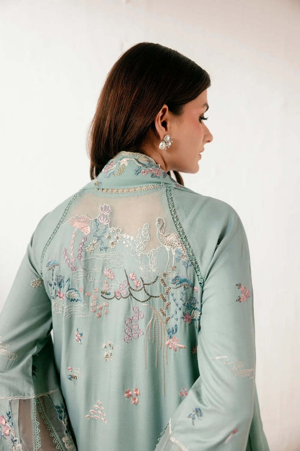 A woman shown from the back wearing a light sage green embroidered traditional garment with floral and bird motifs in pink, blue, and gold, against a plain off-white background.