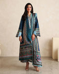 A woman modeling a long teal and navy printed kurta with colorful geometric borders, paired with striped trousers and clear high-heeled sandals, standing against a beige textured wall.
