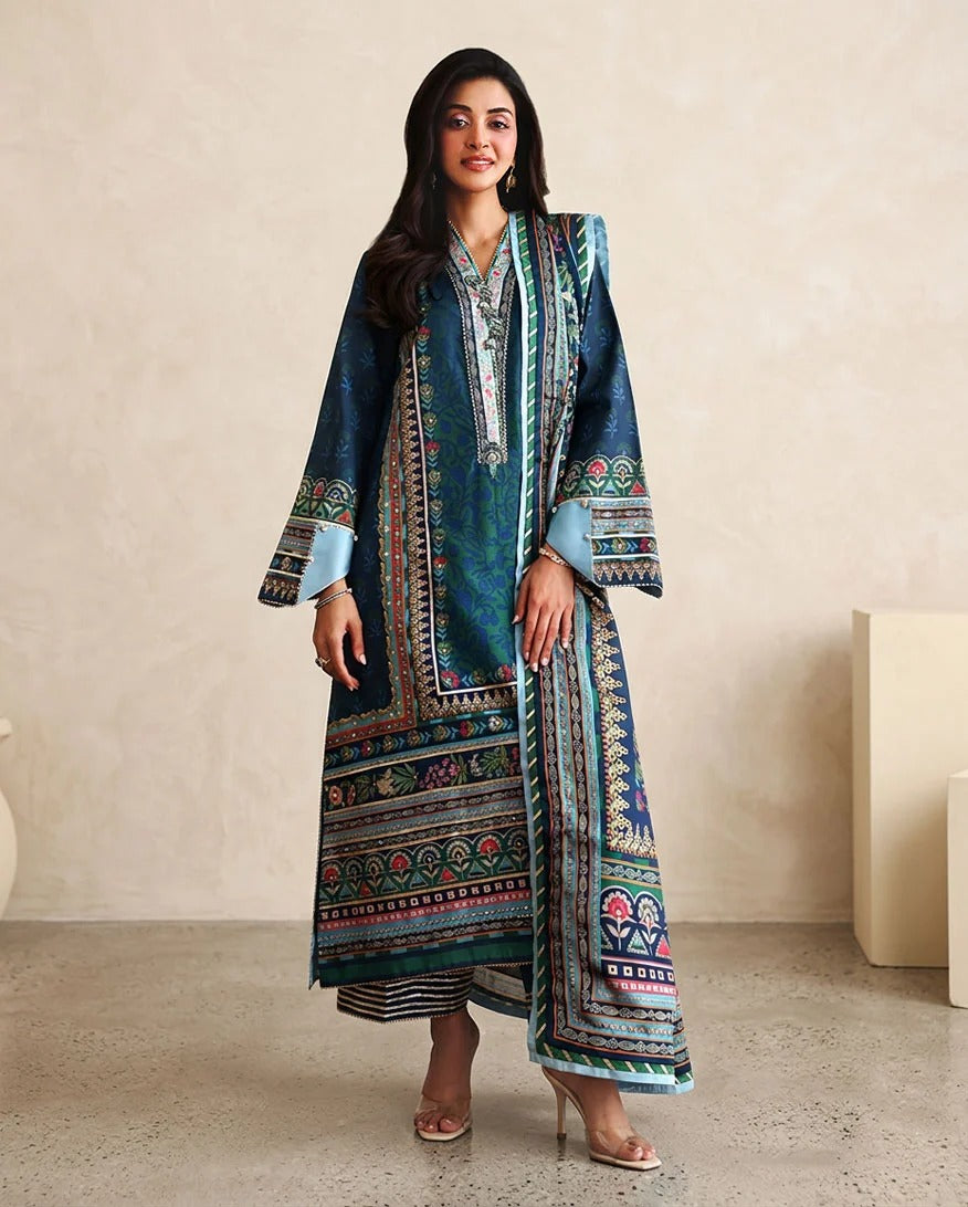 A woman modeling a long teal and navy printed kurta with colorful geometric borders, paired with striped trousers and clear high-heeled sandals, standing against a beige textured wall.