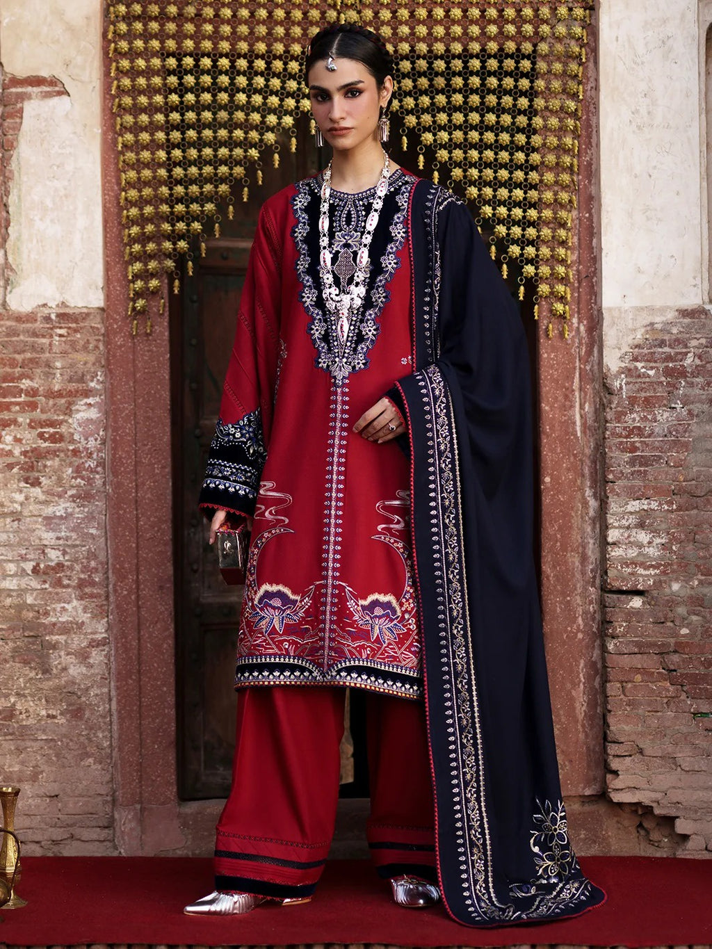 A woman stands in front of an ornate doorway wearing a traditional South Asian outfit. The outfit consists of a deep red kurta and matching wide-leg trousers with elaborate silver, black, and gold embroidery, paired with a dark navy blue dupatta featuring a wide embroidered border.