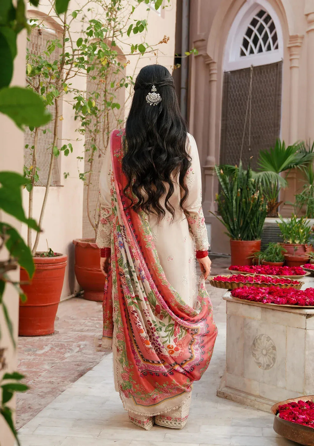 Woman in a traditional outfit standing in a courtyard with plants and flowers.