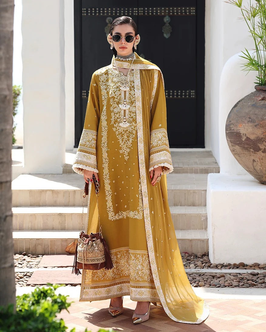 A woman stands outdoors in front of a white building doorway wearing a long golden yellow embroidered traditional outfit with a matching sheer draped overlay, white embellished borders, and holding a small brown handbag.