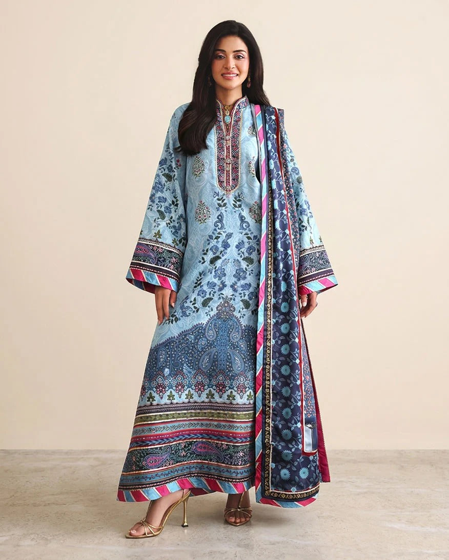 A woman modeling a traditional light blue three-piece salwar kameez suit with intricate multicolor floral prints and borders, paired with a matching dupatta, against a solid beige studio background.