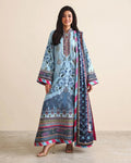 A woman modeling a traditional light blue three-piece salwar kameez suit with intricate multicolor floral prints and borders, paired with a matching dupatta, against a solid beige studio background.