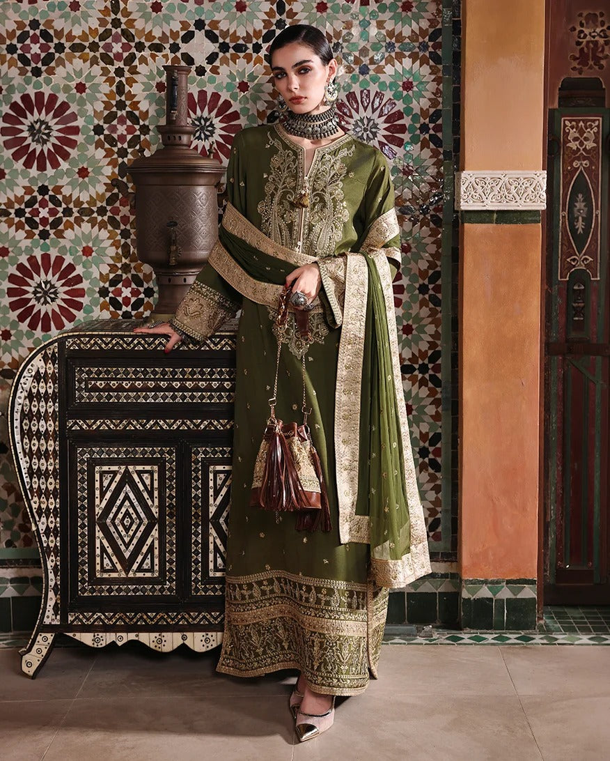 A woman models an olive green traditional South Asian outfit with intricate gold embroidery, a matching dupatta, and a small tassel bag, posed in front of a highly decorative mosaic tile wall and wooden furniture.