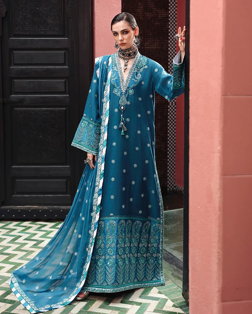 A woman models a traditional teal blue Anarkali suit with white embroidery, a matching dupatta, and a long embroidered coat, posed in front of a pink wall and dark wooden door.