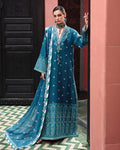 A woman models a traditional teal blue Anarkali suit with white embroidery, a matching dupatta, and a long embroidered coat, posed in front of a pink wall and dark wooden door.