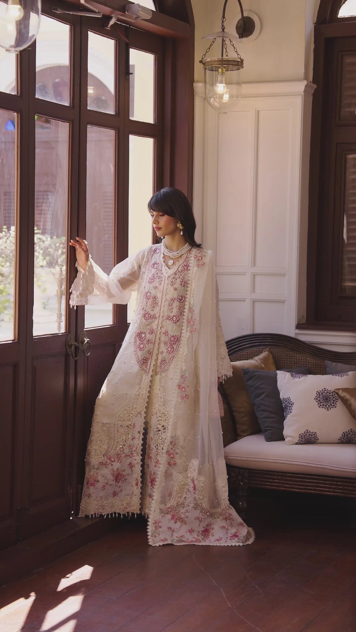 A woman models a traditional white embroidered kurta set with pink floral patterns and a matching dupatta in a sunlit room with dark wood doors and a cushioned bench.