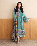 A woman models a traditional three-piece green salwar kameez suit with purple and white printed patterns, standing against a beige wall and wooden paneling.