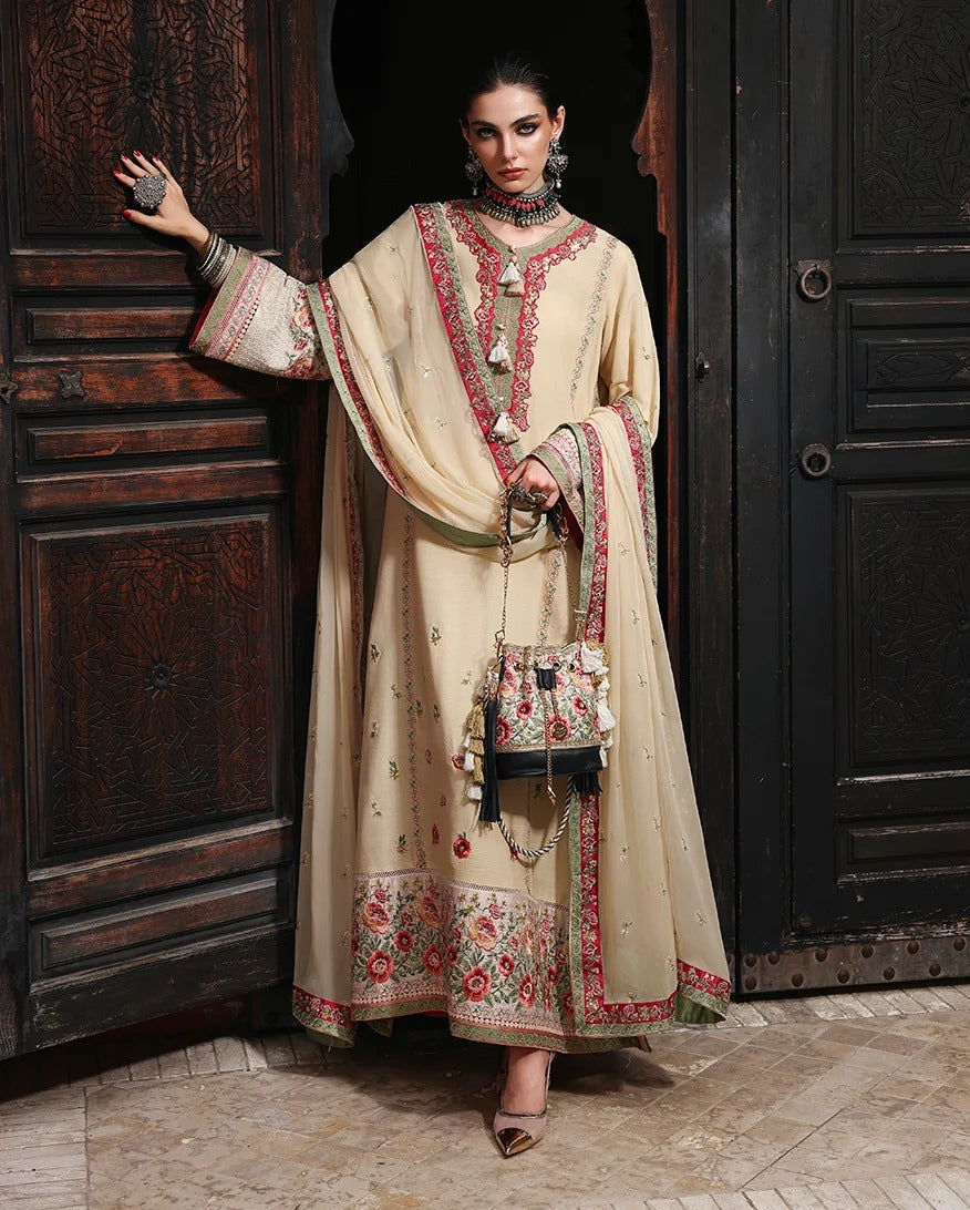 A woman models a traditional beige embroidered outfit with red and green floral detailing, holding a matching embroidered handbag in front of dark carved wooden doors.