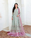 A woman models a traditional Indian anarkali gown with a matching dupatta in a light sage green and dusty pink colorway, heavily embellished with silver embroidery, set against a white architectural background.