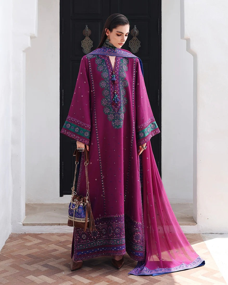 A woman models a traditional magenta purple ethnic outfit with intricate teal and gold embroidery, a matching long dupatta, and a small brown handbag, standing in front of a dark doorway.