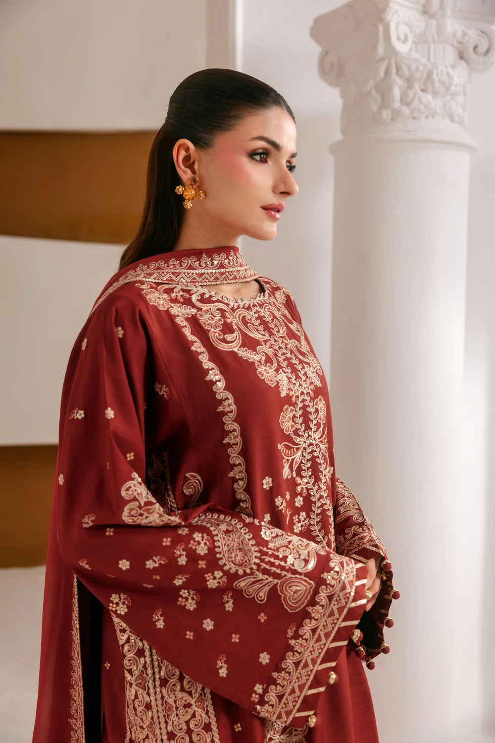 A woman wearing a deep maroon traditional embroidered outfit with intricate gold floral patterns, standing in a bright interior with white columns.