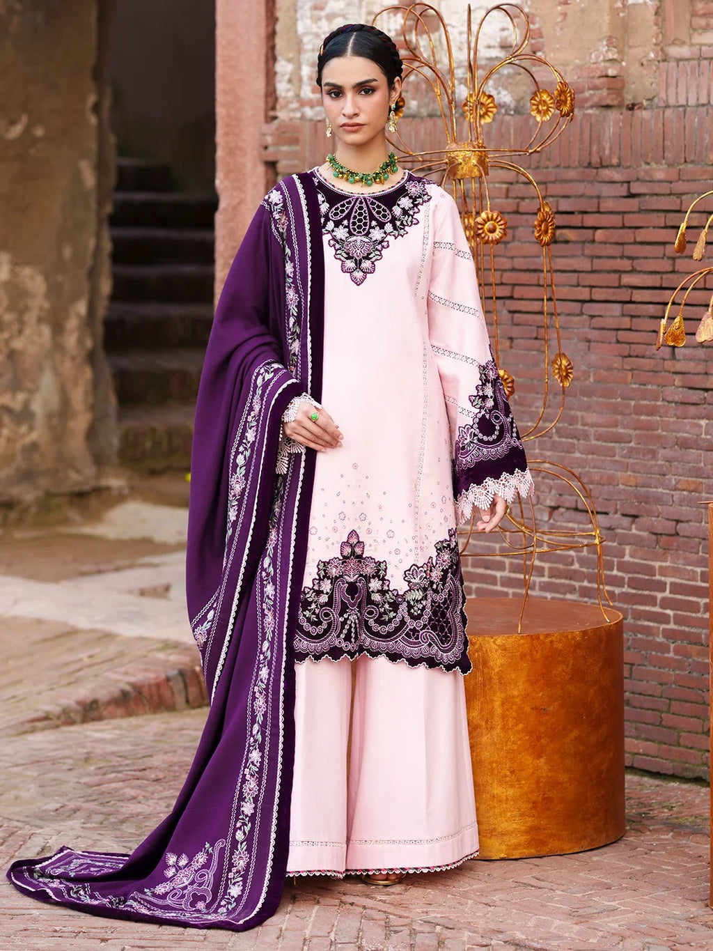 A woman models a traditional South Asian outfit consisting of a light pink kurta with dark purple embroidery, matching wide-leg trousers, and a dark purple dupatta with white embroidery, set against a rustic brick wall background.