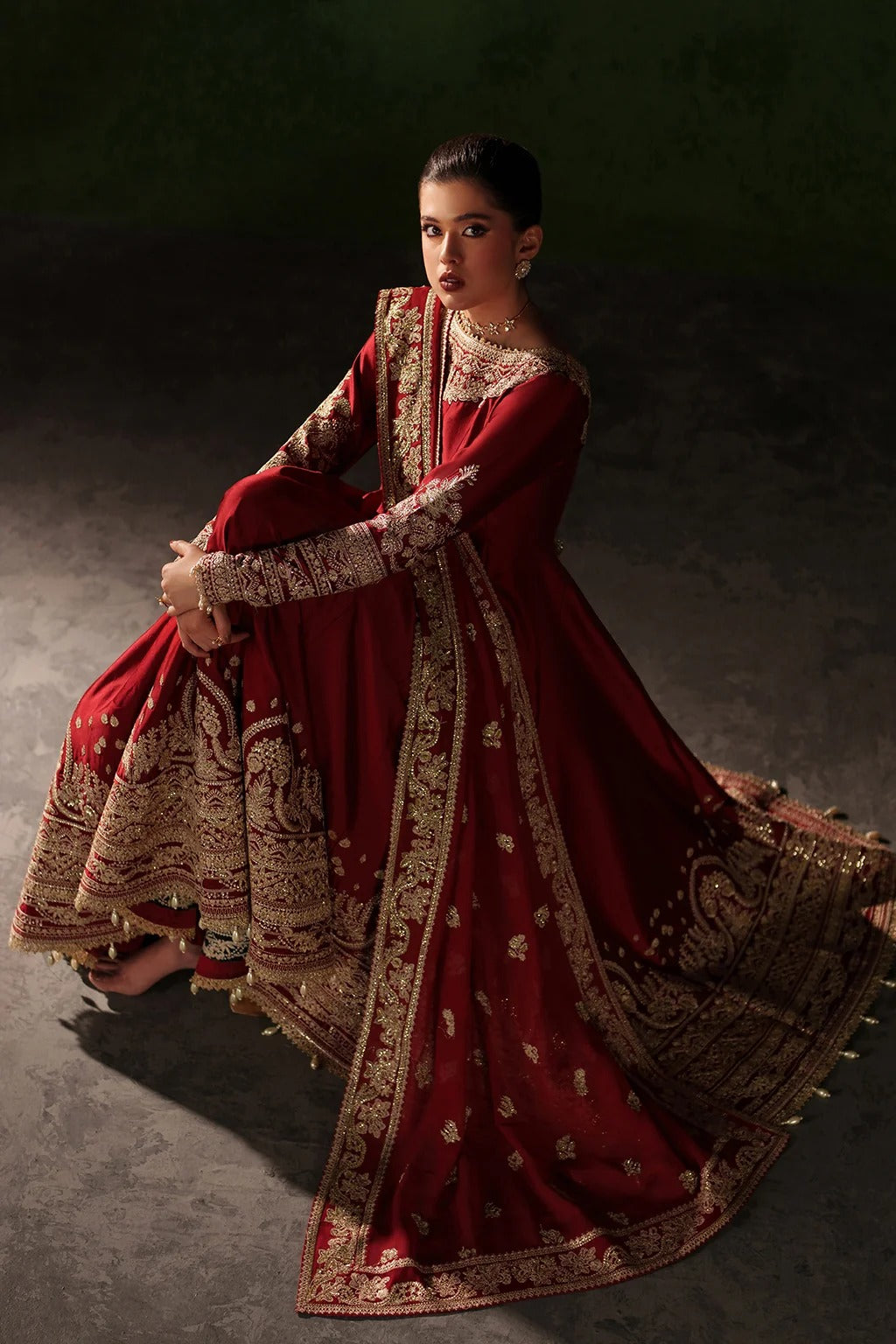 A woman wearing a traditional red and gold embroidered Anarkali gown, seated in a dramatic studio setting with a dark green background.