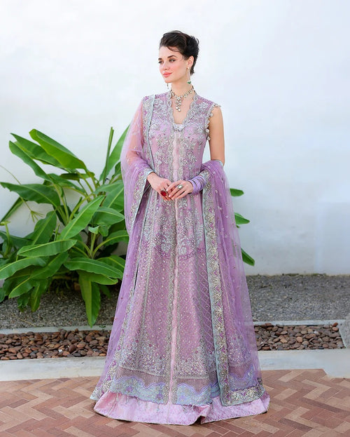 A woman models a lavender purple embroidered traditional Anarkali gown with a matching dupatta, standing outdoors against a white wall with green plants.