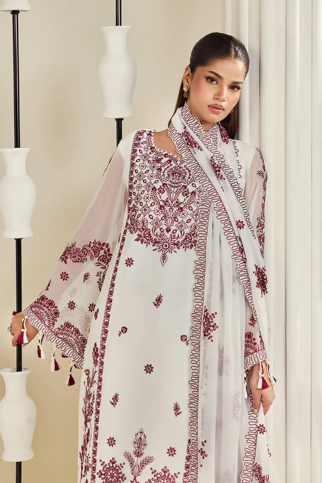 A woman models a traditional white and maroon embroidered kurta set with a matching dupatta, standing in front of a neutral background with decorative white vases.