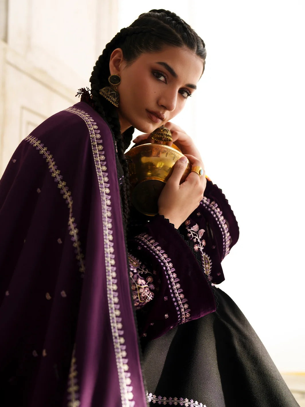 A woman wearing a deep purple embroidered traditional outfit with a matching draped shawl and black skirt, holding a gold-toned metal pot. She is posed indoors against a bright white background.