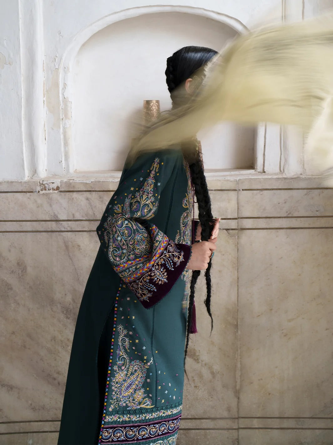 A woman in profile wearing a dark teal embroidered traditional outfit with intricate multicolor patterns and a long flowing beige scarf, standing against a beige stone wall.
