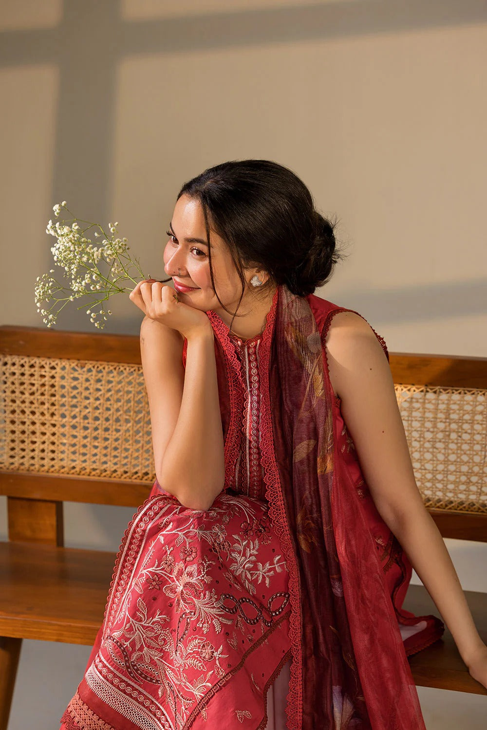 A woman wearing a red embroidered traditional outfit with a matching sheer dupatta, seated on a wooden bench with a cane backrest against a beige wall.