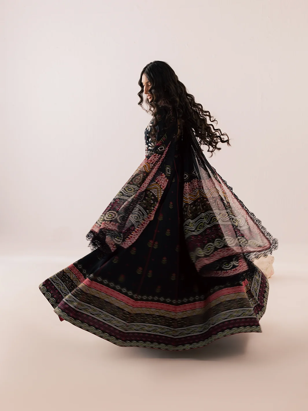A woman wearing a black embroidered traditional dress with pink and beige patterned dupatta and flared skirt, captured mid-spin against a plain beige studio background.