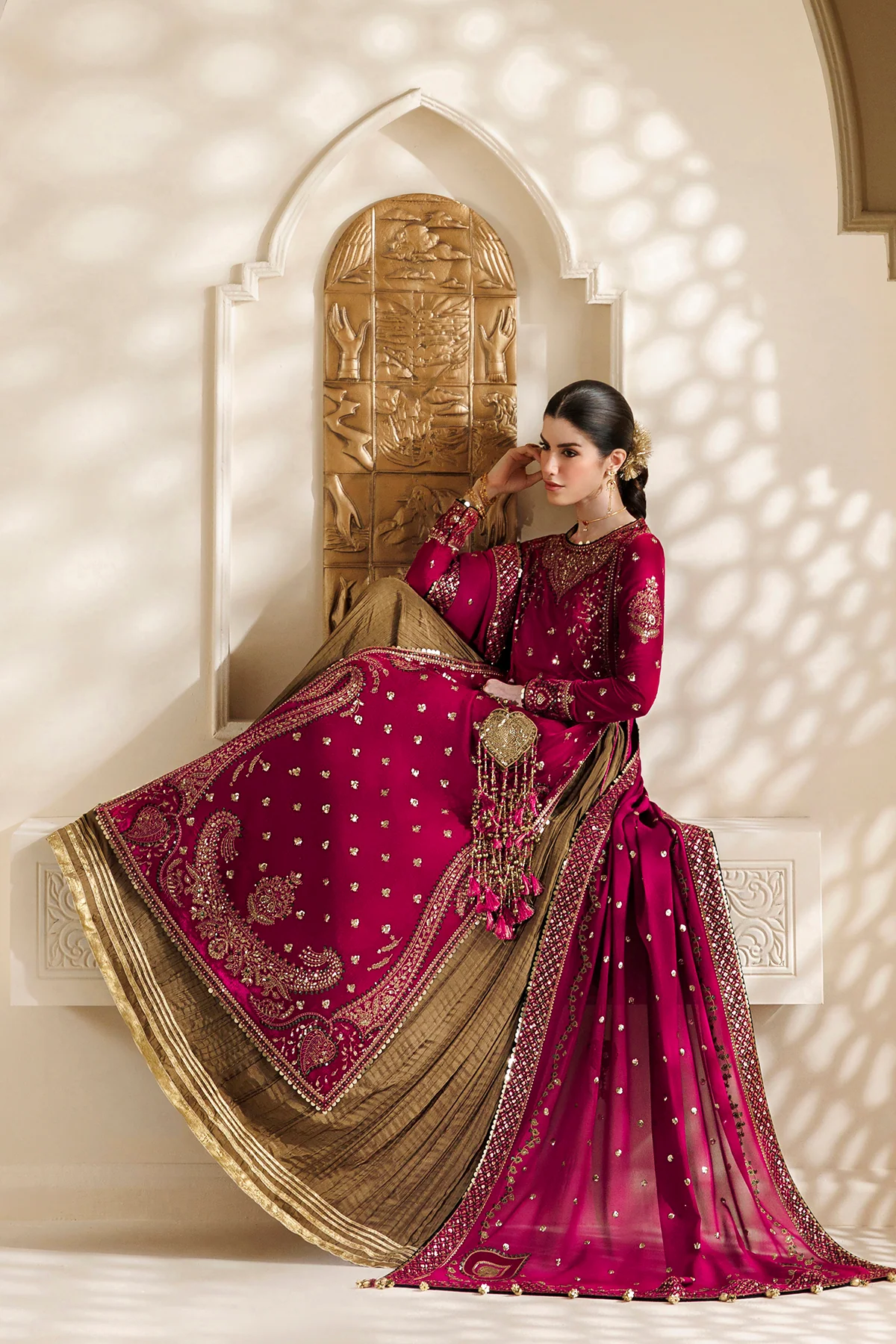A woman models an ornate magenta and gold traditional outfit with a heavily embroidered dupatta, posed in front of a carved beige stone archway with soft shadow patterns.
